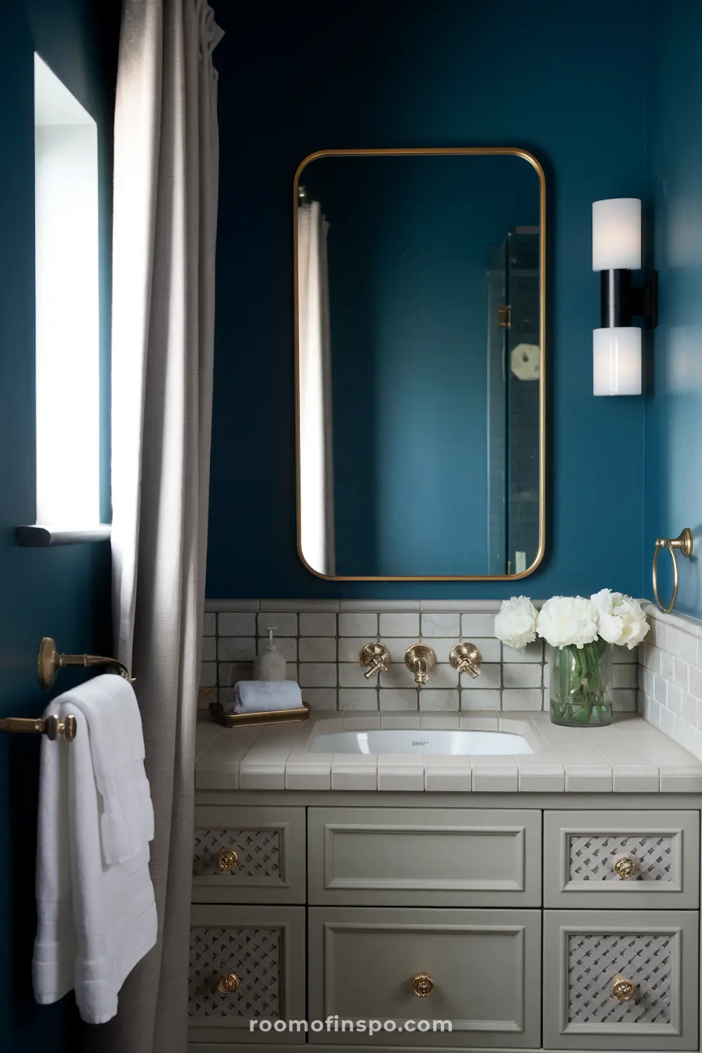 Um banheiro estiloso com paredes azul-escuras, um grande espelho retangular com moldura dourada, uma pia branca com acessórios dourados, uma bancada com azulejos brancos, um vaso de flores brancas e uma luminária com duas lâmpadas cilíndricas.