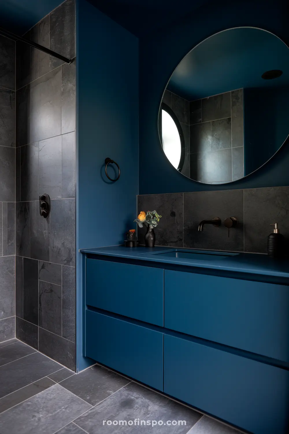 Um banheiro moderno com armários azul escuro, um grande espelho redondo, uma torneira preta e uma área de chuveiro com azulejos cinza escuro.