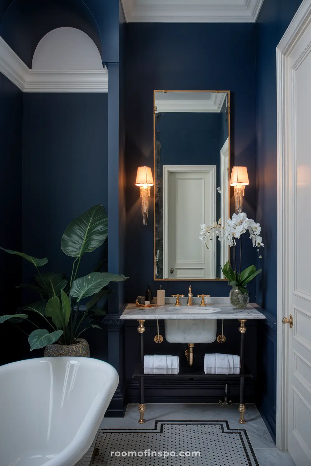 Um banheiro luxuoso com paredes azul-escuras, um grande espelho com borda dourada, dois apliques de parede com cúpulas, uma pia de mármore com torneiras douradas, uma planta em vaso, um vaso com orquídeas brancas, e uma banheira branca.