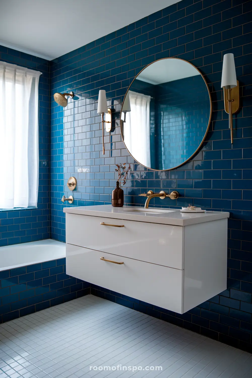 Um banheiro moderno com paredes de azulejos azul em forma de ladrilho, um espelho redondo, uma vaidade flutuante branca com acessórios dourados, uma torneira de parede, e dois apliques de parede, apresentando uma janela com cortinas brancas e uma banheira.