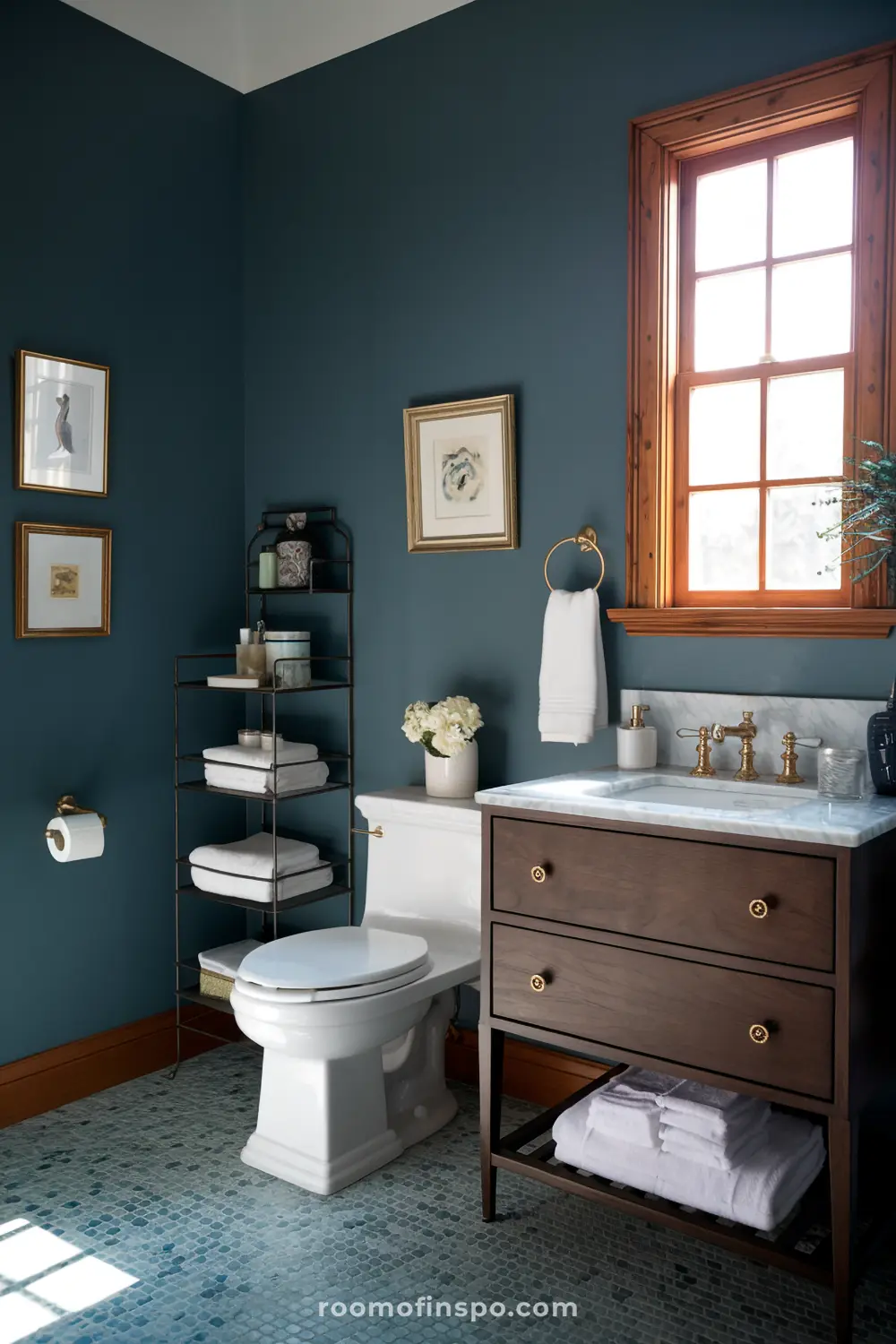 Um banheiro pequeno e bem organizado com paredes azul-escuras, um vaso sanitário branco, uma vaidade de madeira com uma bancada de mármore, uma janela com moldura de madeira, uma estante metálica com toalhas e produtos de higiene, e arte emoldurada nas paredes.
