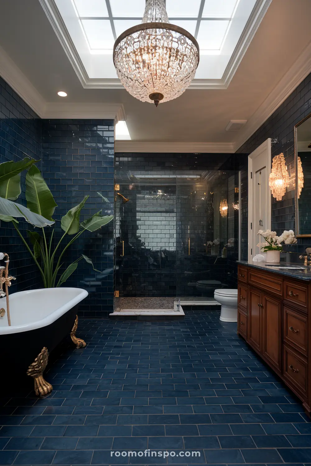 Um banheiro luxuoso com azulejos escuros em forma de ladrilho, uma banheira independente com pés dourados, uma planta grande, uma vaidade de madeira com uma bancada de mármore, um vaso sanitário, um chuveiro envidraçado, e um candelabro pendurado em uma claraboia.