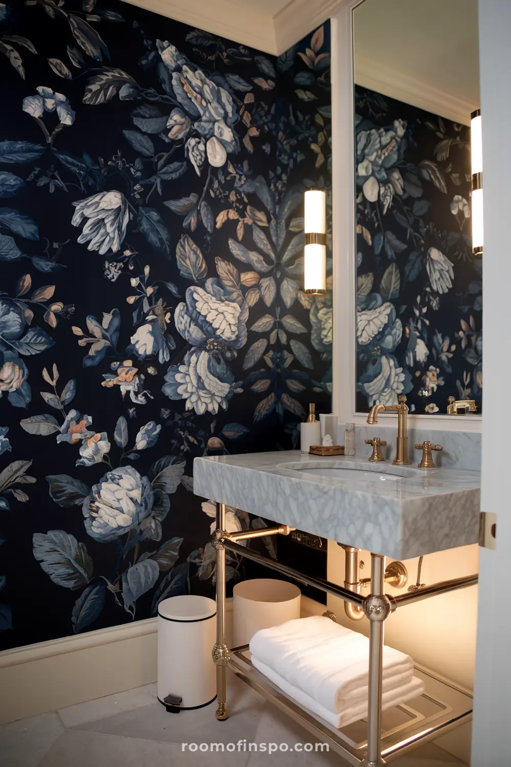 Um banheiro com papel de parede floral azul escuro, uma vaidade de mármore e acessórios de bronze com iluminação suave.