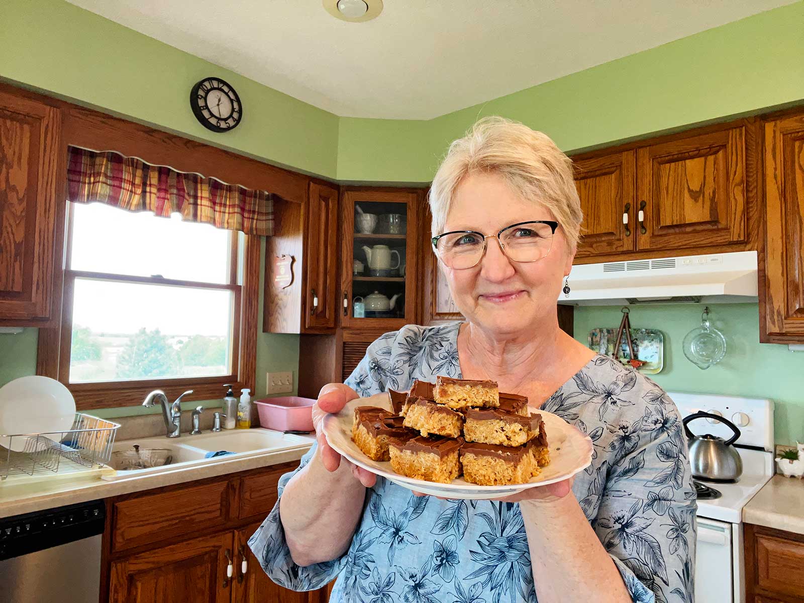 Nana sorrindo e segurando um prato de scotcharoos em sua cozinha.