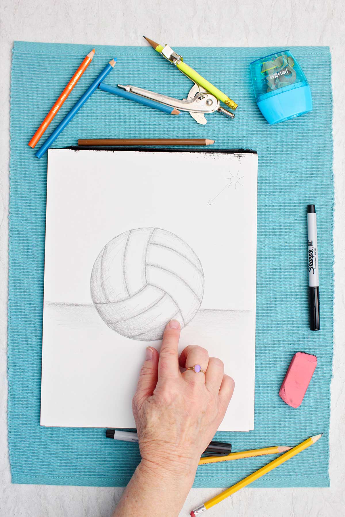 Desenho de uma bola de vôlei a lápis com uma pessoa sombreando com o dedo e materiais ao lado em um lugar de mesa azul.