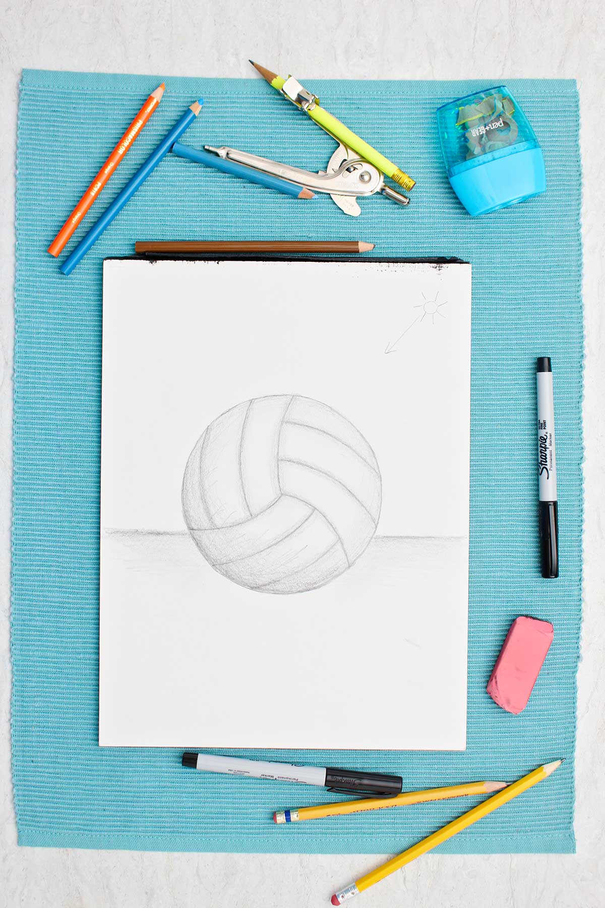 Desenho de uma bola de vôlei a lápis com sombreamento e materiais ao lado em um lugar de mesa azul.