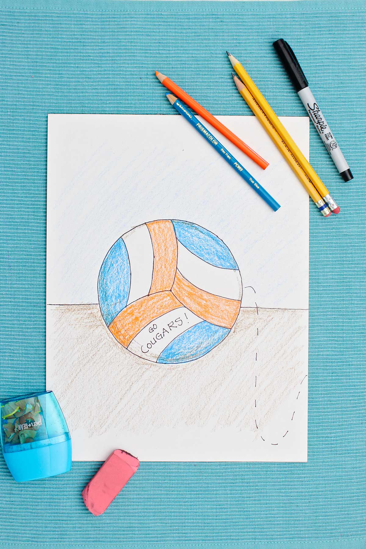 Desenho de vôlei colorido com lápis de cor azul e laranja com materiais próximos em um jogo americano azul.