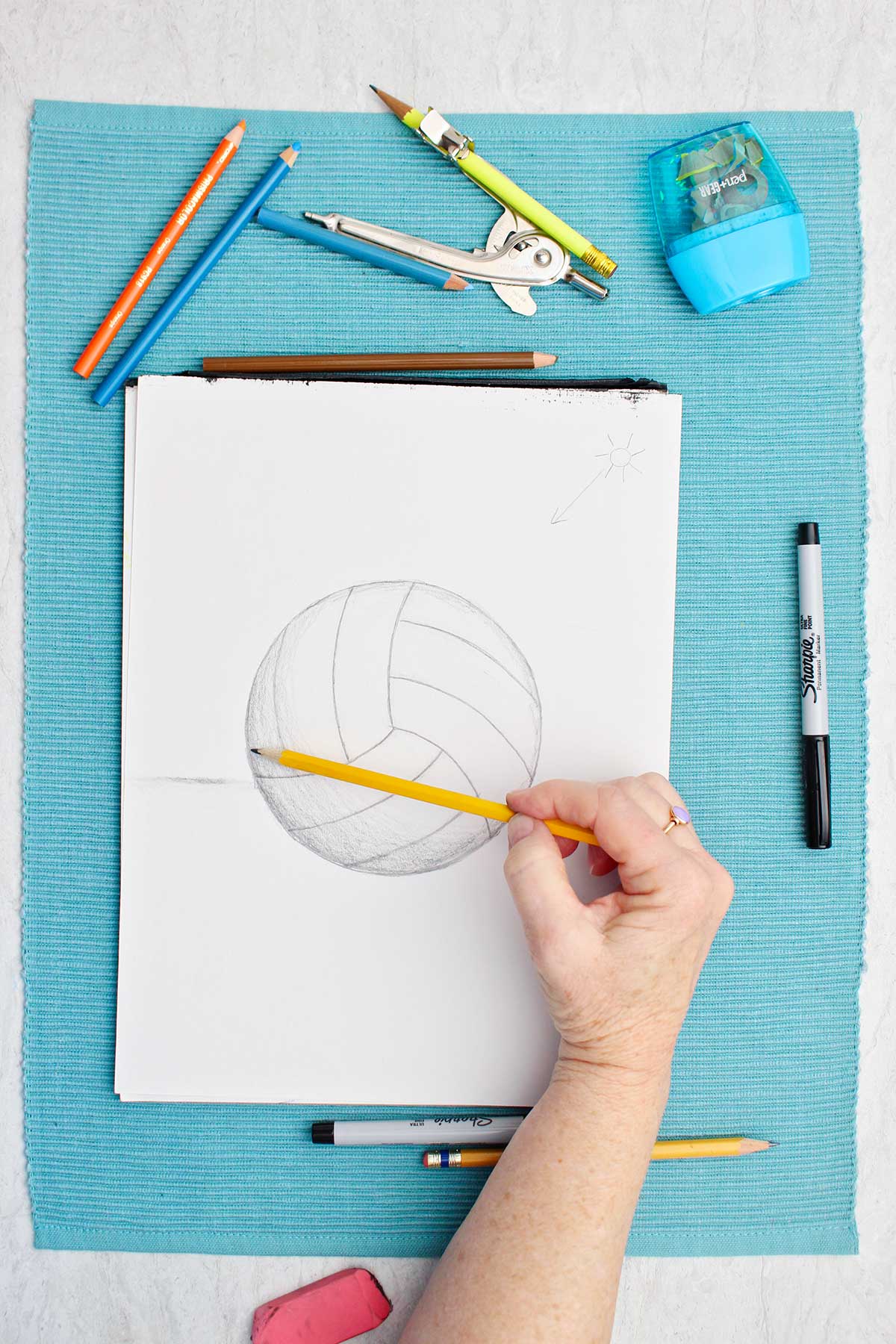 Desenho de uma bola de vôlei a lápis com uma pessoa sombreando com o dedo e materiais ao lado em um lugar de mesa azul.