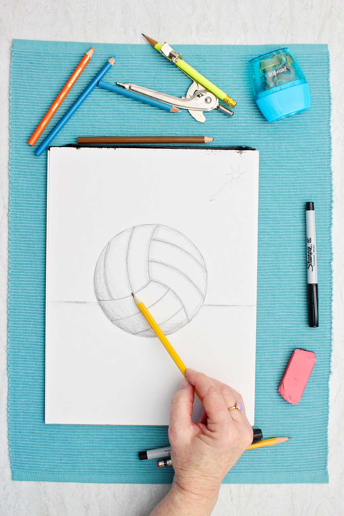 Desenho de uma bola de vôlei a lápis com uma pessoa sombreando com lápis e materiais ao lado em um lugar de mesa azul.