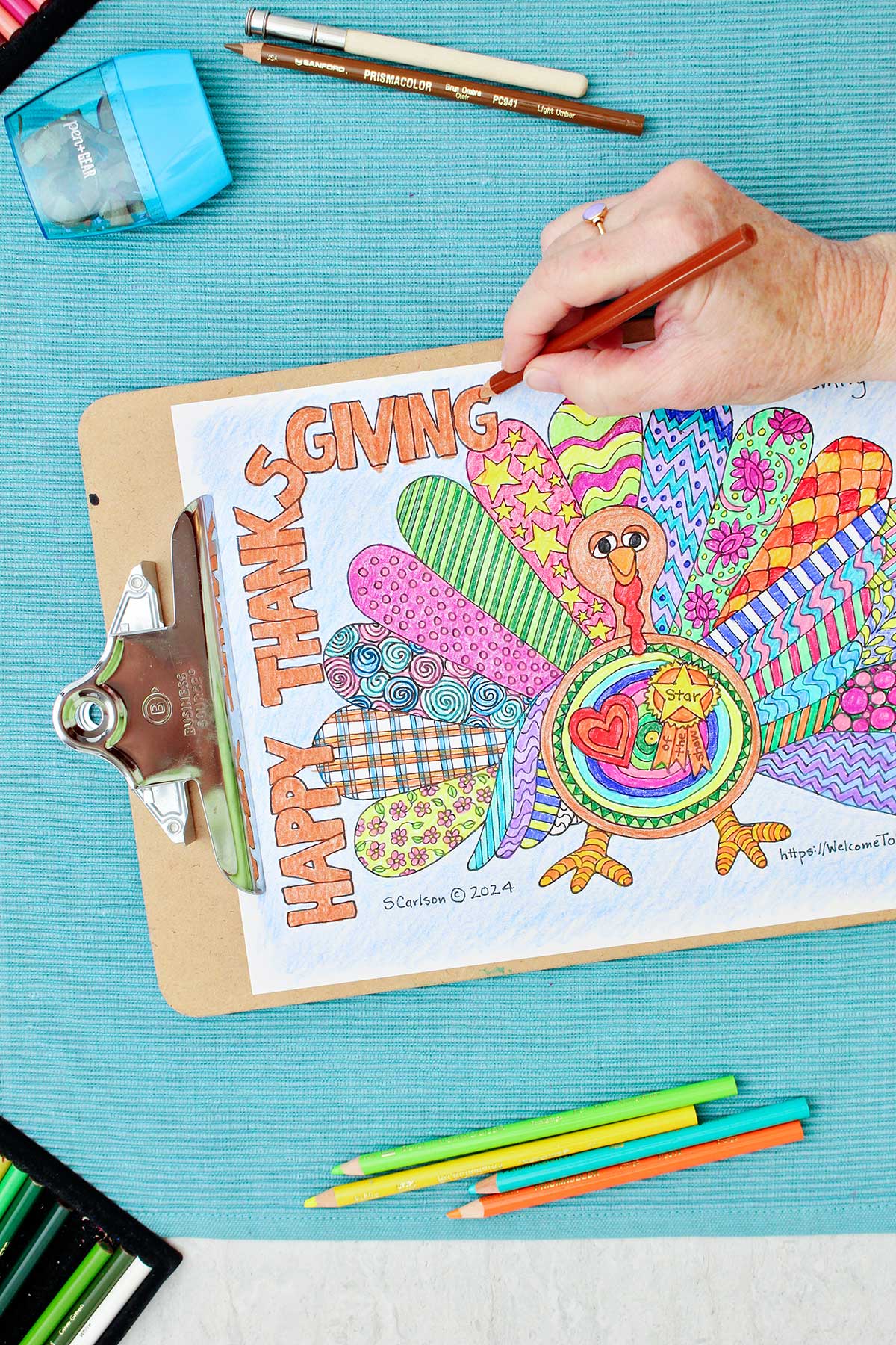 Página de colorir de Ação de Graças de um peru com asas padronizadas está presa a um prancheta e repousa sobre um lugar-manto aqua com lápis de cor próximos. Pessoa sombreadando o rosto do peru em marrom.