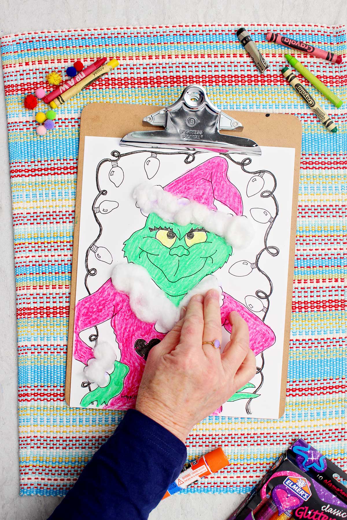 Pessoa pressionando bolas de algodão nos detalhes do casaco do Papai Noel na página de colorir do Grinch.