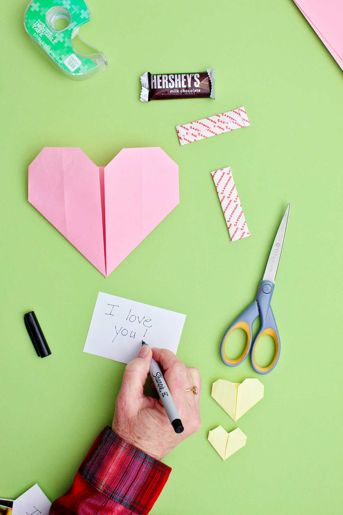 Uma pessoa escrevendo uma nota dizendo "Eu te amo!" para colocar em um coração de origami rosa.