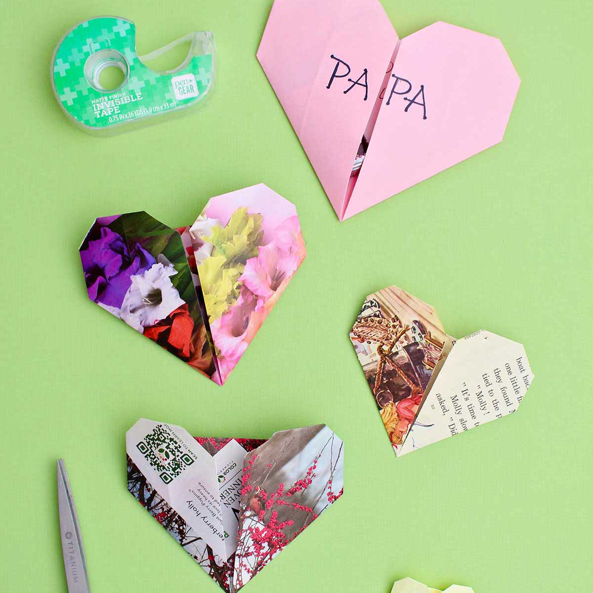 Quatro corações de origami em um fundo verde com tesoura e fita próximas.