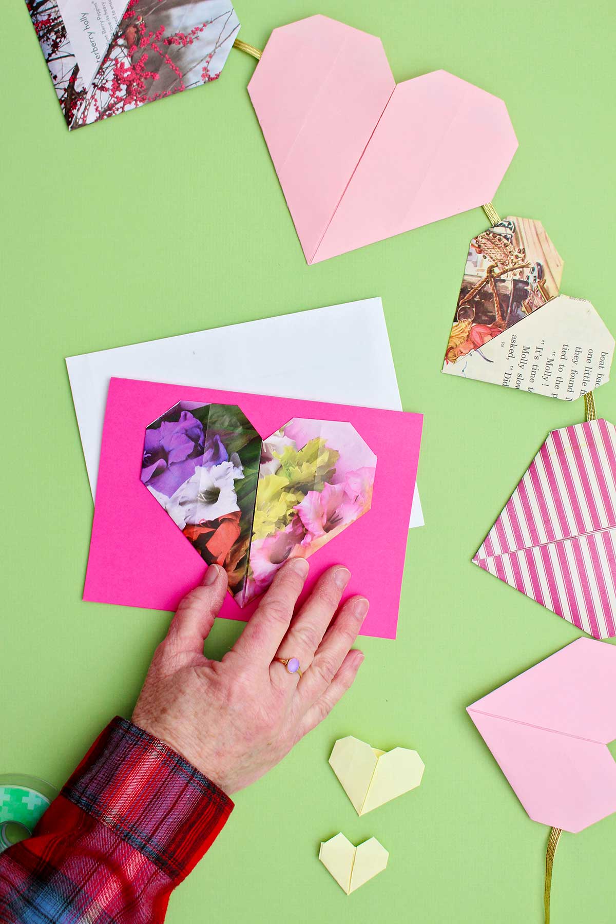 Um coração de origami floral sendo colocado em um cartão rosa com outros corações de origami próximos.