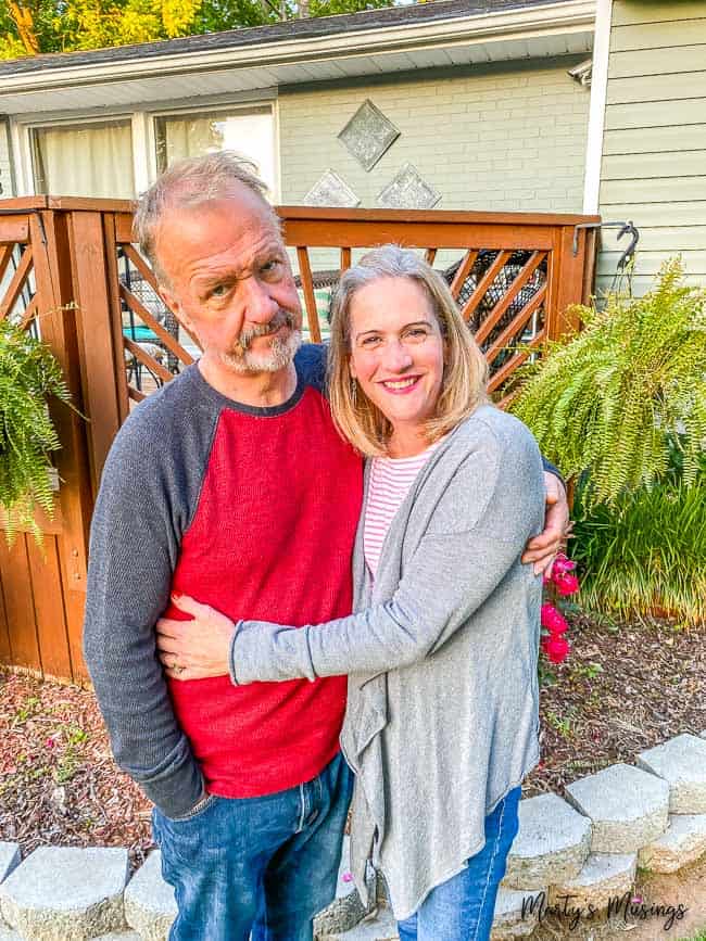 Marido com expressão preocupada enquanto esposa o abraça na frente da casa