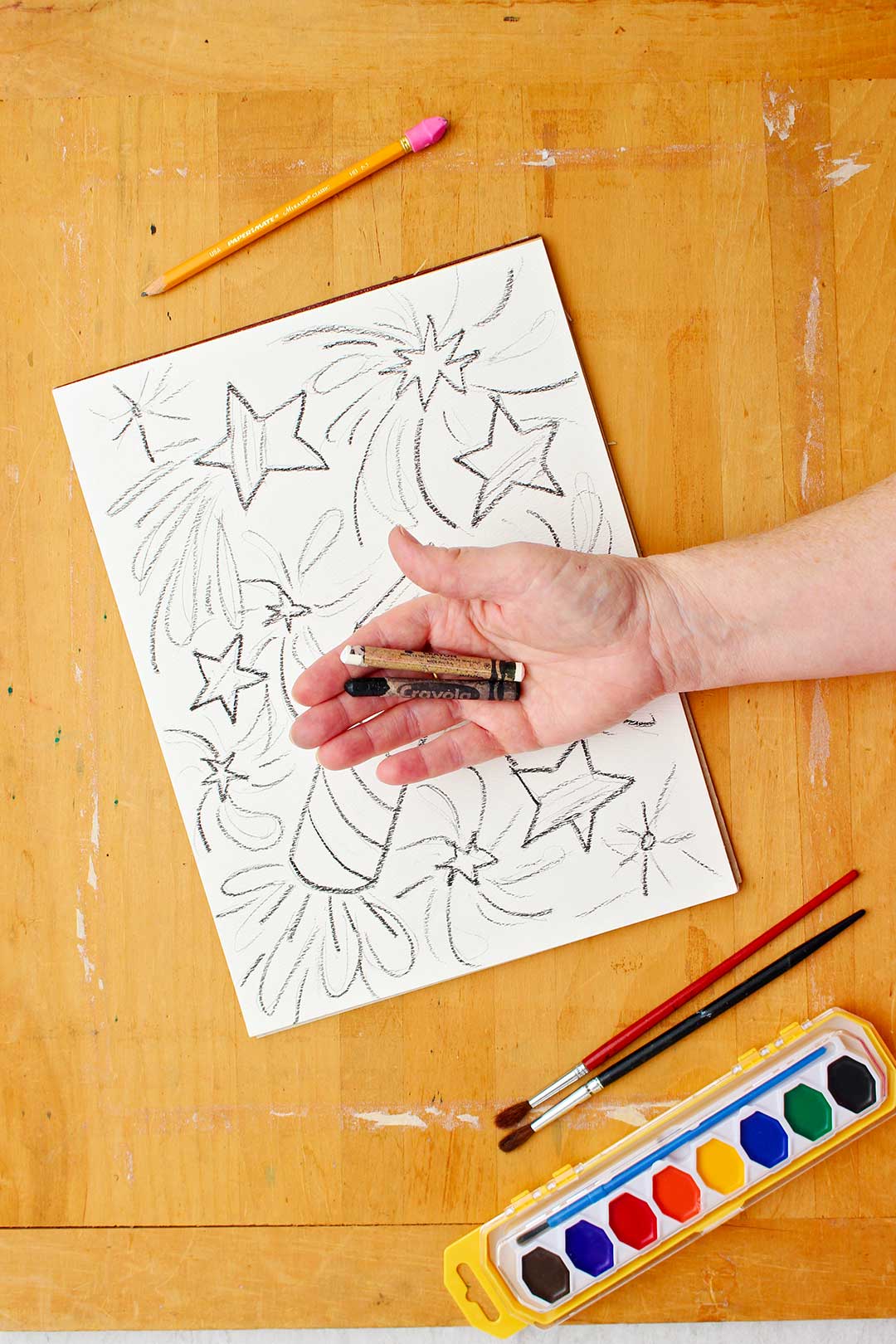 Mão segurando um lápis de cera preto e branco acima de um desenho de estrelas e foguetes.