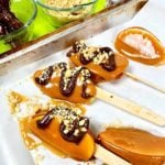 Fatias de maçã caramelizada concluídas no palito descansando sobre papel manteiga. Algumas têm espirais de chocolate amargo e nozes polvilhadas.