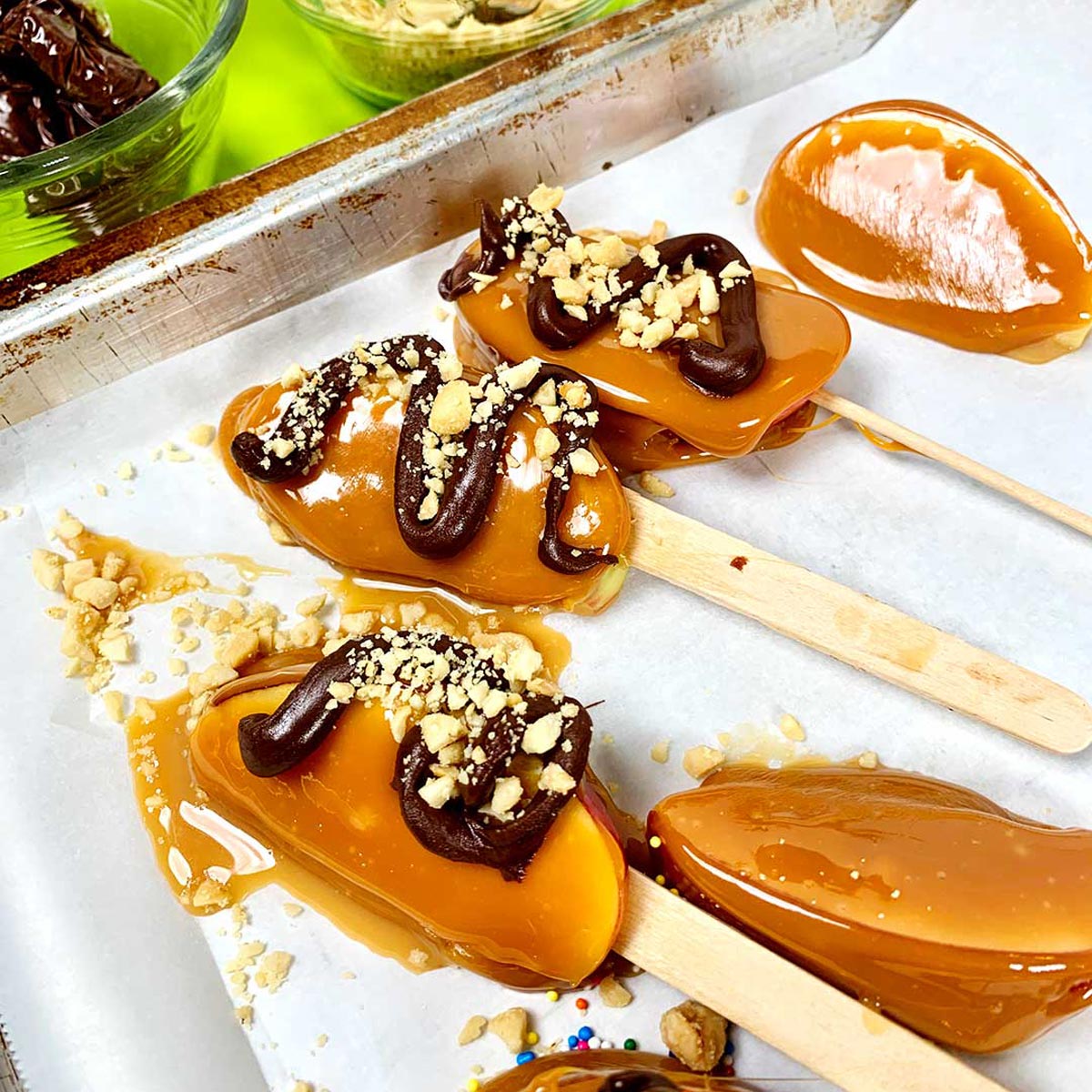Fatias de maçã caramelizada completas em palitos repousando sobre papel manteiga. Algumas têm espirais de chocolate meio amargo e nozes polvilhadas.