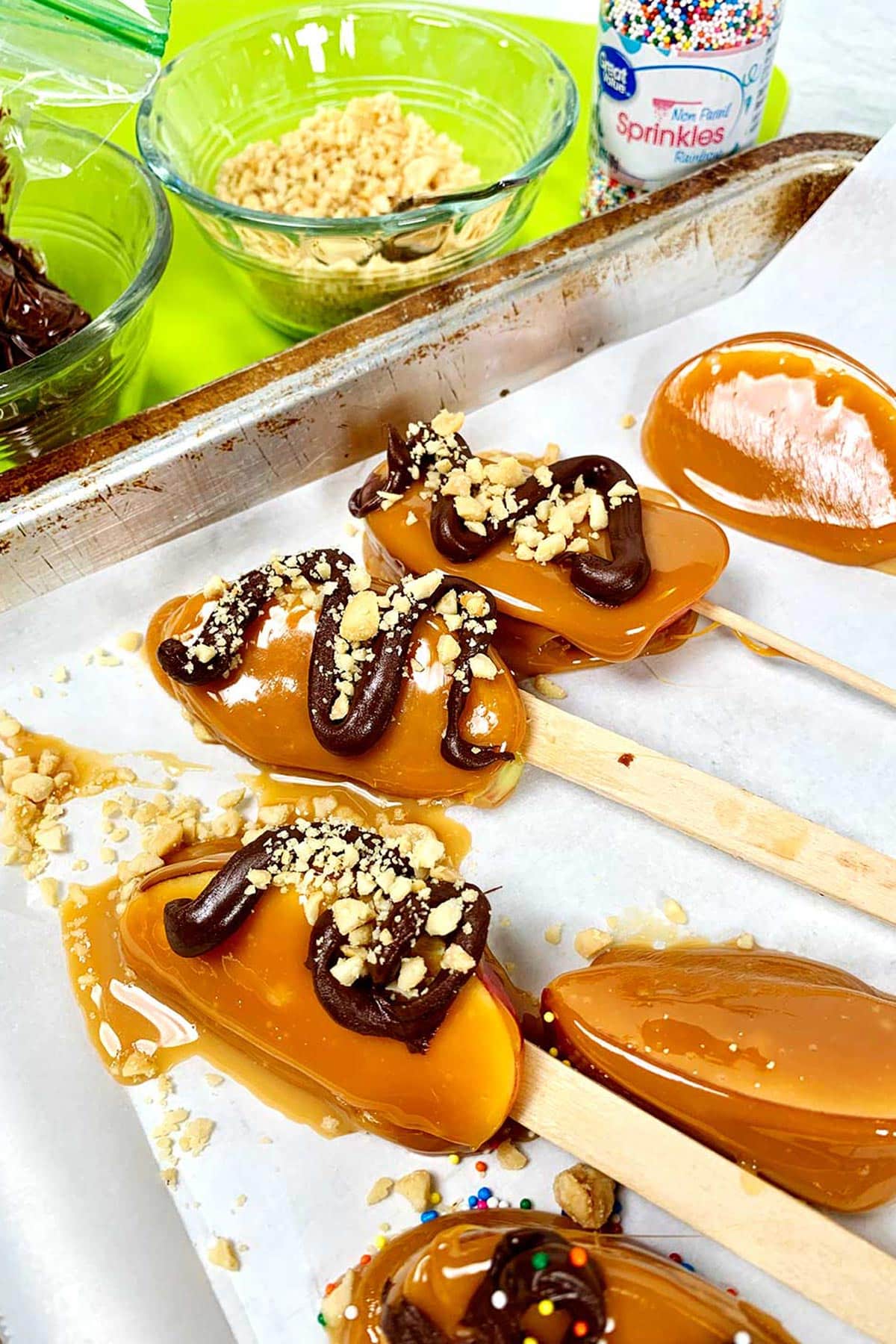 Fatias de maçã caramelizada finalizadas no palito repousando sobre papel manteiga. Algumas têm espirais de chocolate amargo e nozes polvilhadas.