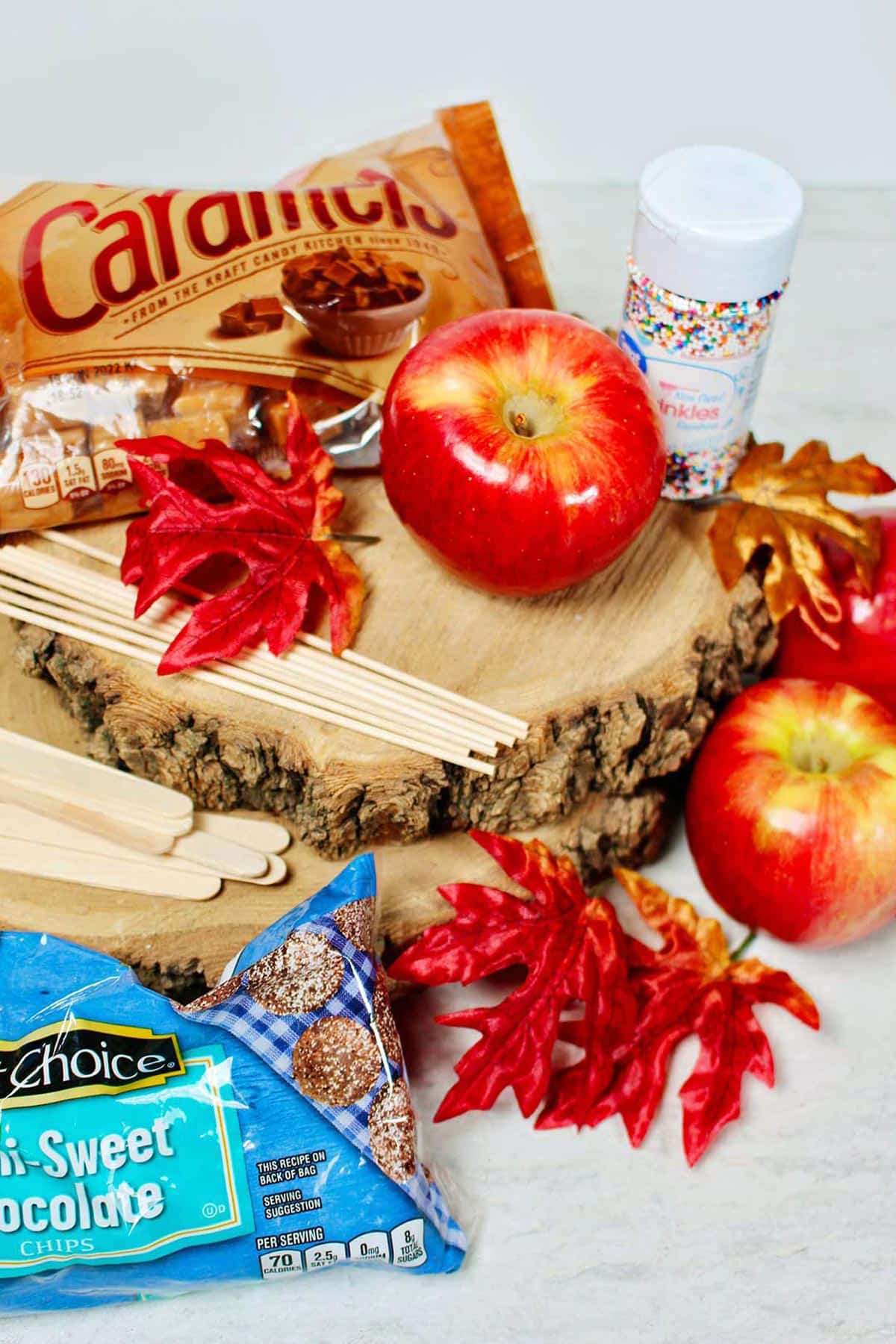 Exibição da temporada de outono com maçãs vermelhas, palitos, gotas de chocolate meio amargo, caramelos, granulados e folhas sobre fatias de madeira.