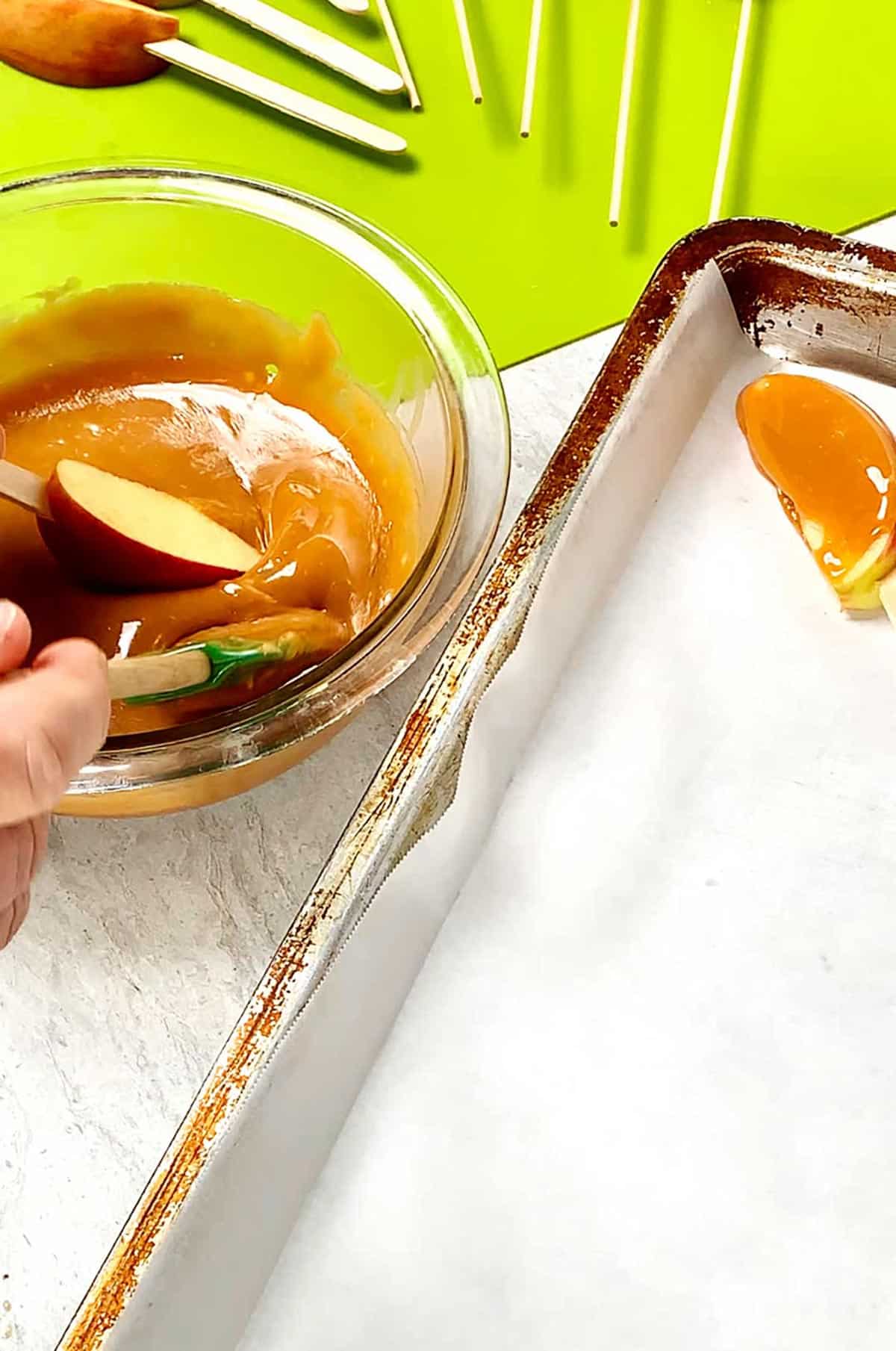 Pessoa mergulhando fatias de maçã no palito em caramelo aquecido em uma tigela ao lado de uma tábua de cortar verde brilhante e uma assadeira com papel manteiga.