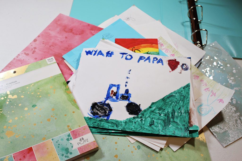 Pintura de uma criança de um trator e uma colina, endereçada ao Papai, papel de scrapbook e um fichário ao fundo.