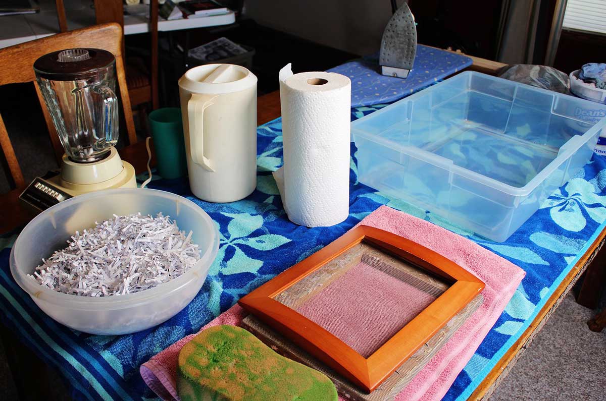Uma tigela de recortes de papel, uma moldura para quadro com molde e deckle, toalhas de papel, uma toalha de praia, um recipiente com água, uma imagem e um ferro.