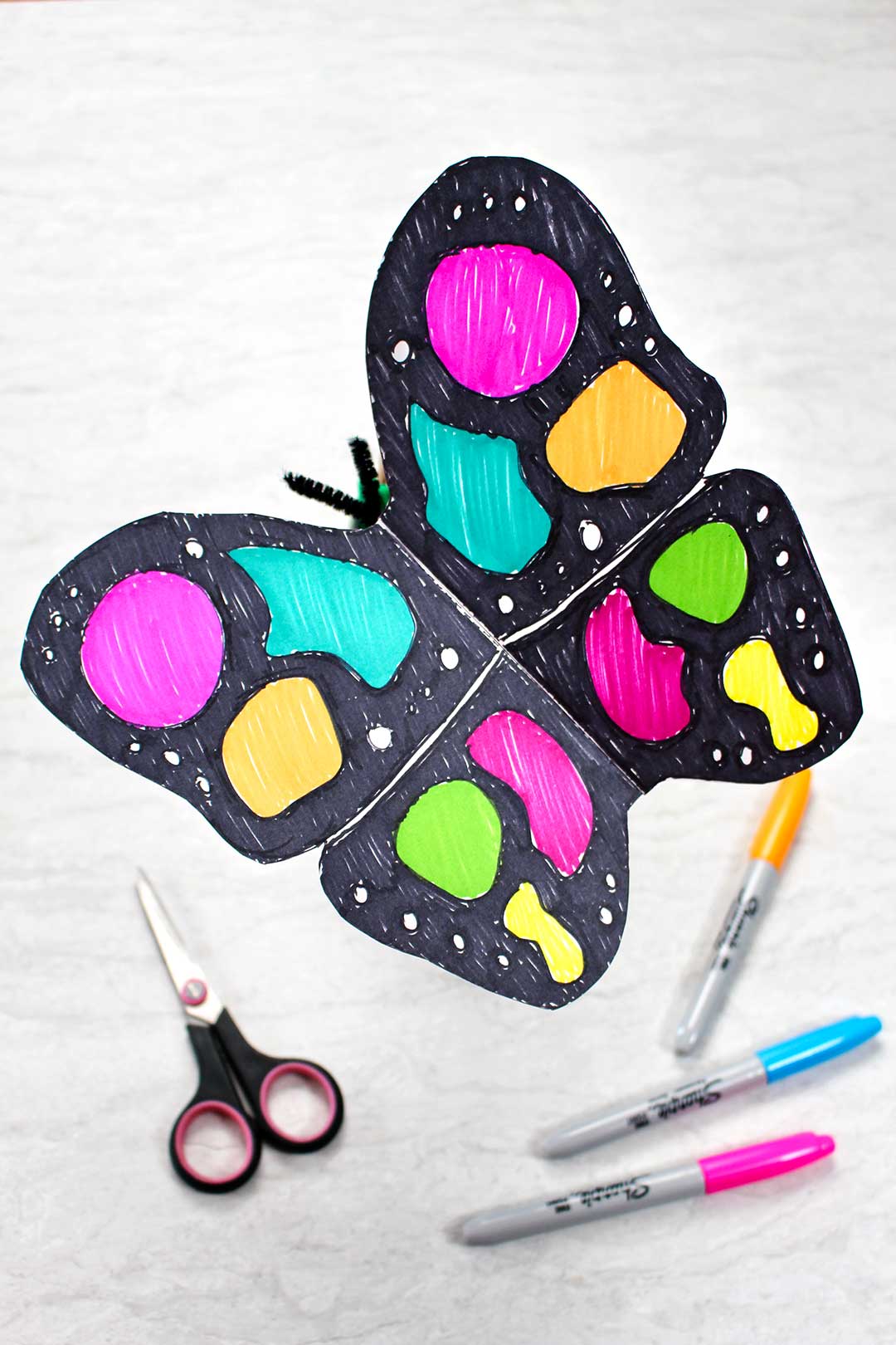 Artesanato de borboleta de papel completo colorido com marcadores preto e neon descansando perto de uma tesoura preta e marcadores neon.
