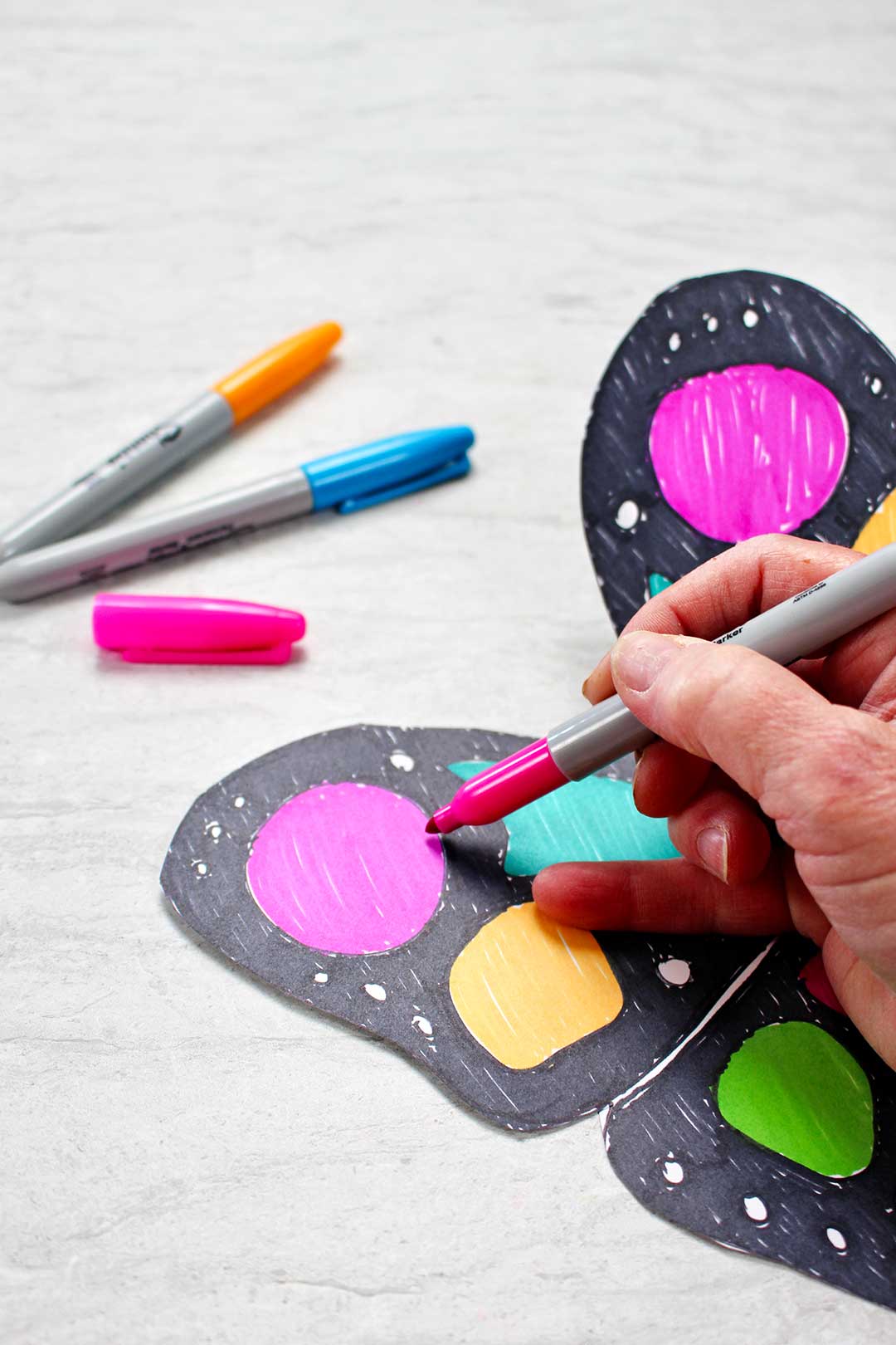 A mão de uma pessoa colorindo uma parte da asa de papel da borboleta com um marcador neon rosa com outros marcadores descansando perto.