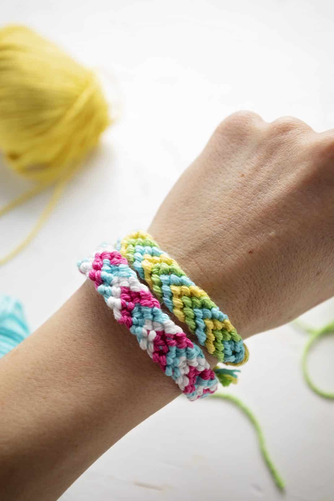 Duas pulseiras de padrão chevron em fio de bordado verde, azul, amarelo, rosa e branco, amarradas no pulso de uma pessoa.