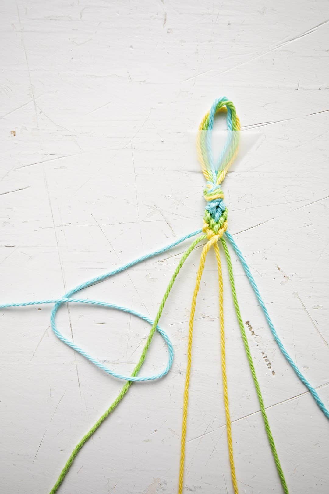 Uma pulseira com padrão Chevron sendo feita com fios azuis, verdes e amarelos.