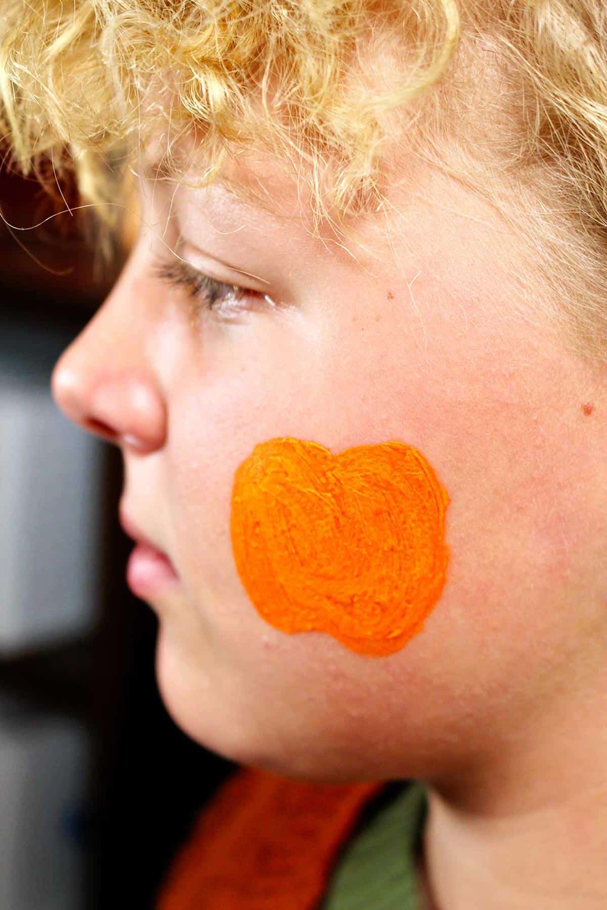 Perfil do rosto de uma garota com contorno de pintura facial laranja de uma abóbora na bochecha.