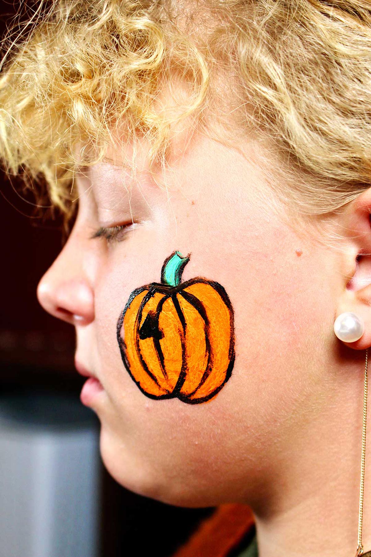 Perfil do rosto de uma garota com pintura mais detalhada de uma abóbora laranja com talo verde e contorno preto.