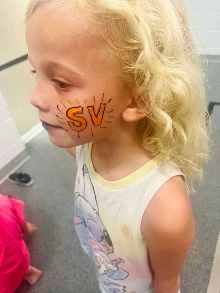 Menina jovem com cabelo loiro com pintura facial laranja "SV".