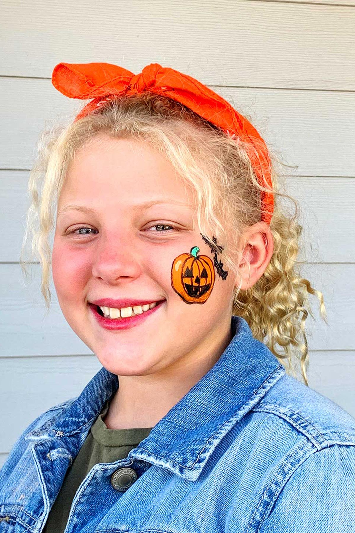 Menina com cabelo cacheado loiro sorrindo para a câmera com pintura de rosto de abóbora de Halloween na bochecha.