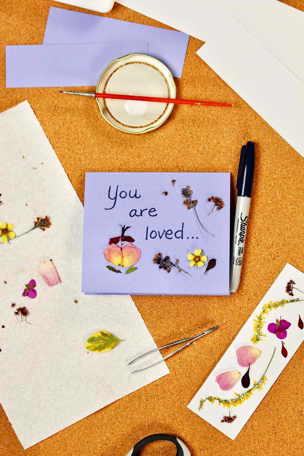 Uma carta roxa dizendo 'Você é amado...' com flores prensadas sobre ela, além de um marcador com flores prensadas.