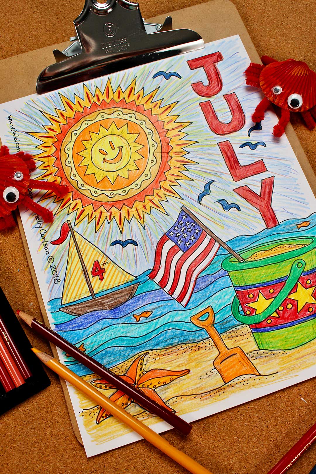 Página de colorir de julho finalizada em prancheta com dois caranguejos DIY e lápis de cor ao redor.