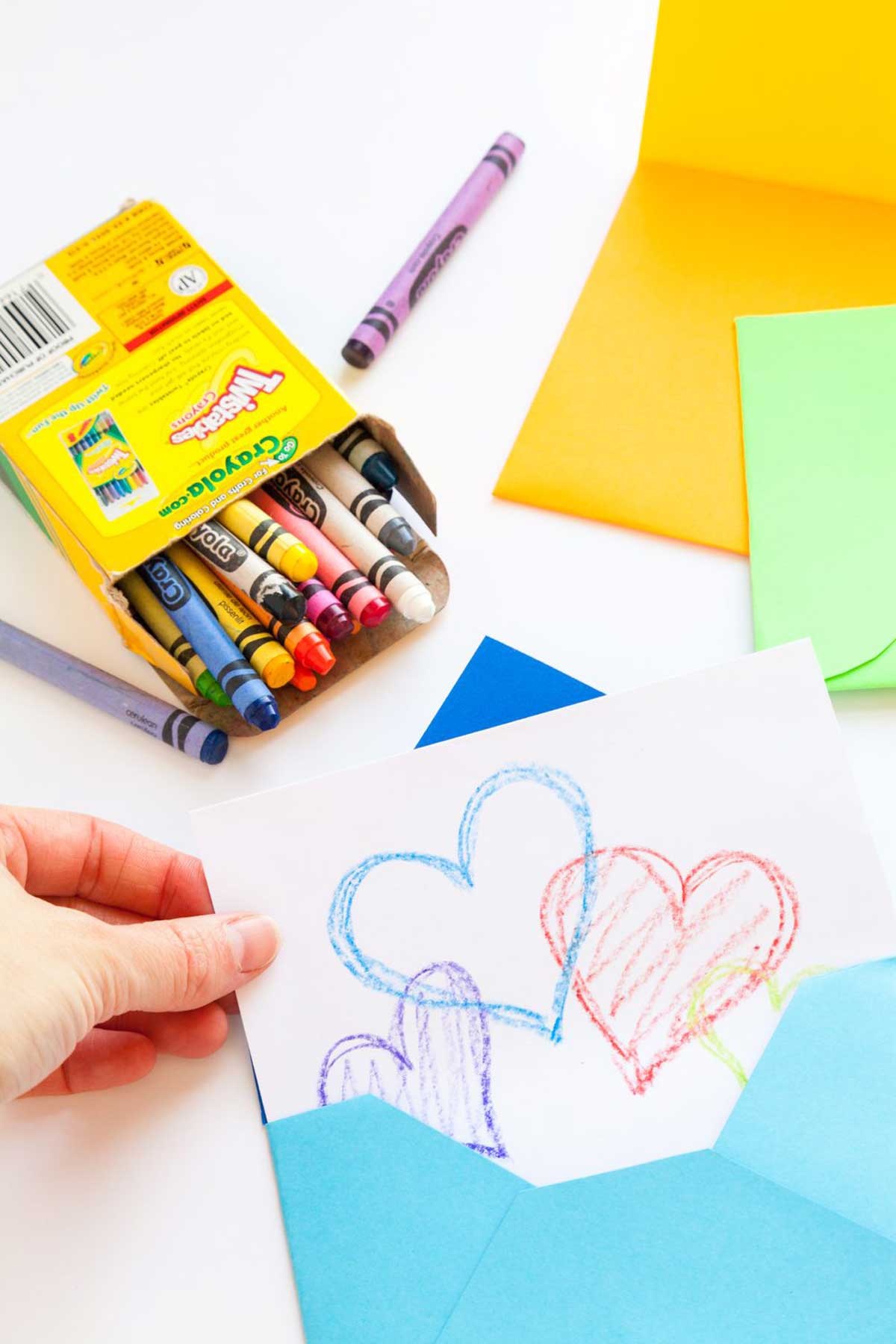 Puxando um cartão com corações de um envelope artesanal azul, com giz de cera e papel colorido ao redor.