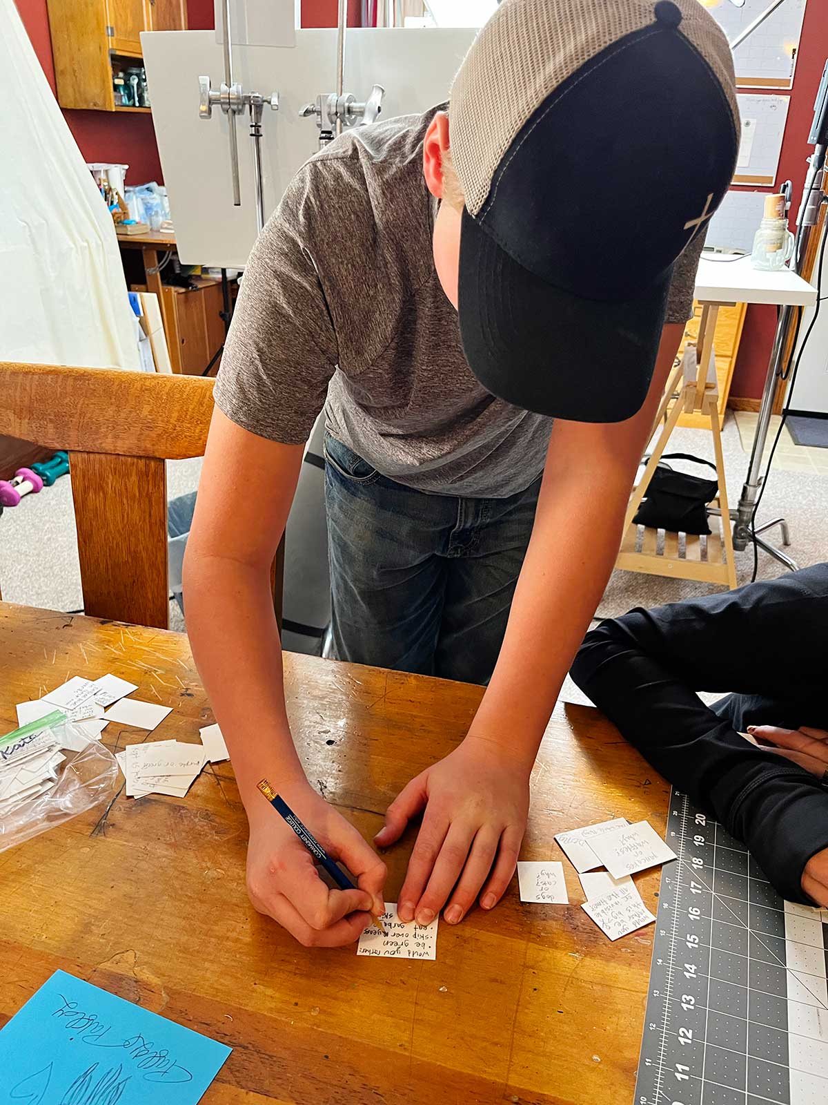 Um menino com um boné de beisebol escrevendo uma pergunta de 'Você Preferiria' em um pedaço de papel.