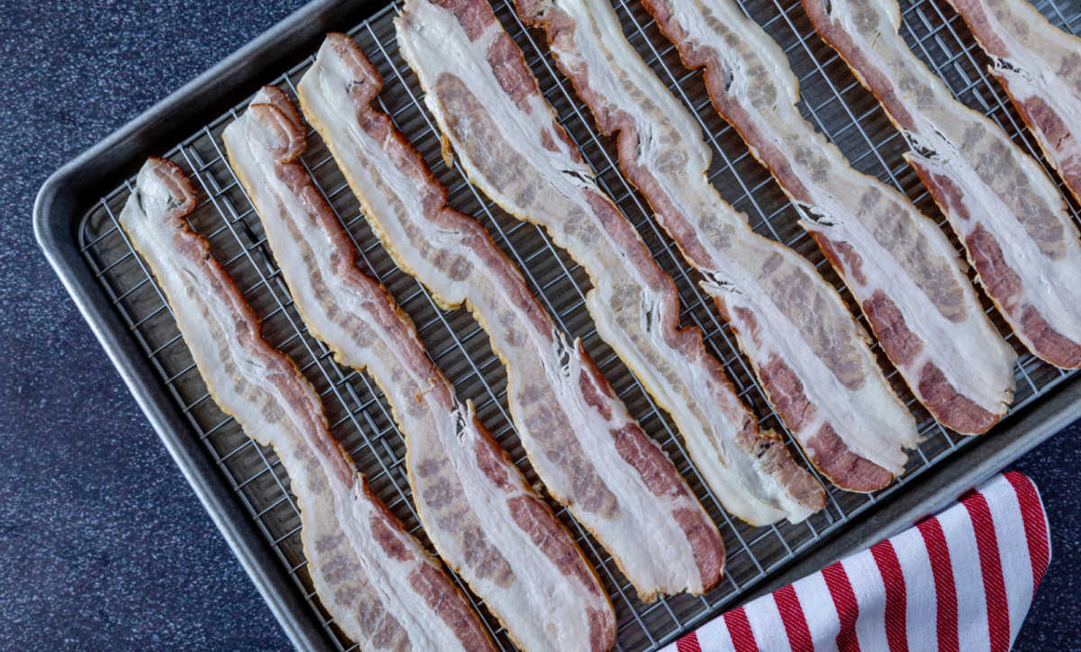 Fatias de bacon em uma grade de arame sobre uma assadeira ao lado de uma toalha vermelha e branca.