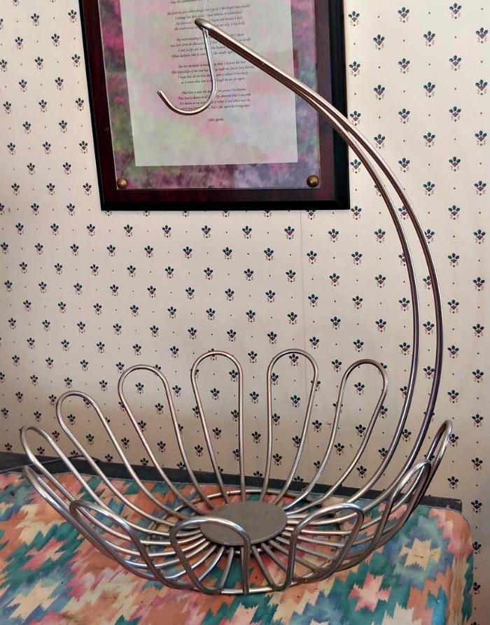Suporte de metal para bananas sobre um caminho em teal e rosa na frente de uma imagem emoldurada com uma citação.