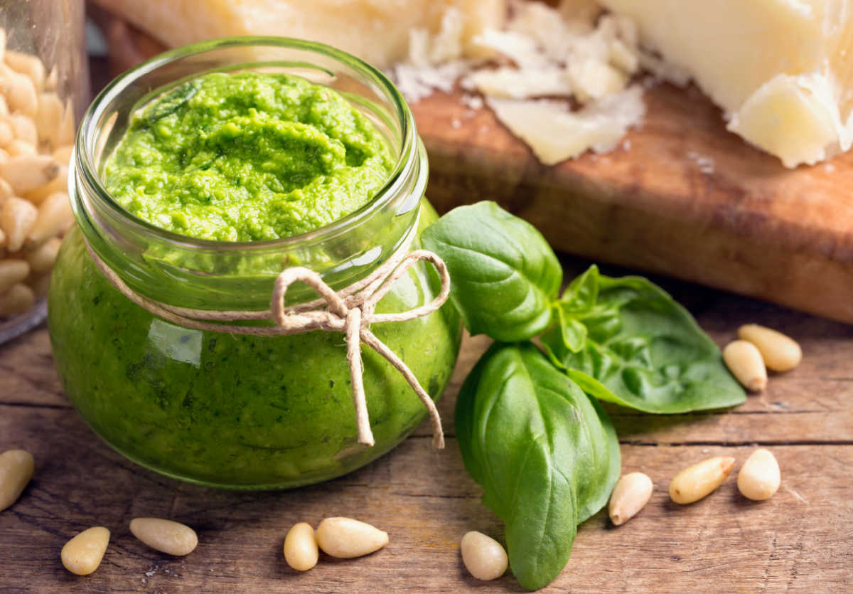 Faça pesto de manjericão Pesto de manjericão em um pote de vidro sobre uma bancada de madeira, ao lado de manjericão fresco, pinhões e queijo parmesão.