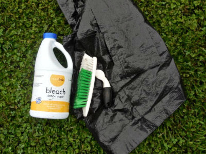 Uma pilha de itens necessários para limpar um bebedouro de pássaros, incluindo um saco de lixo preto de 40 galões, uma escova rígida de esfregar e água sanitária em um gramado. Esses materiais podem ser usados para limpar todos os tipos de bebedouros de pássaros, incluindo bebedouros de concreto, cimento e pedra.