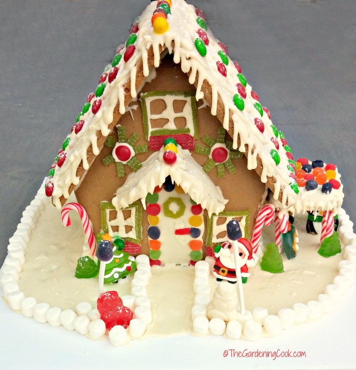 Casa de gengibre com telhado coberto de glacê e enfeites de doces, cercada por um jardim de doces contornado com marshmallows.