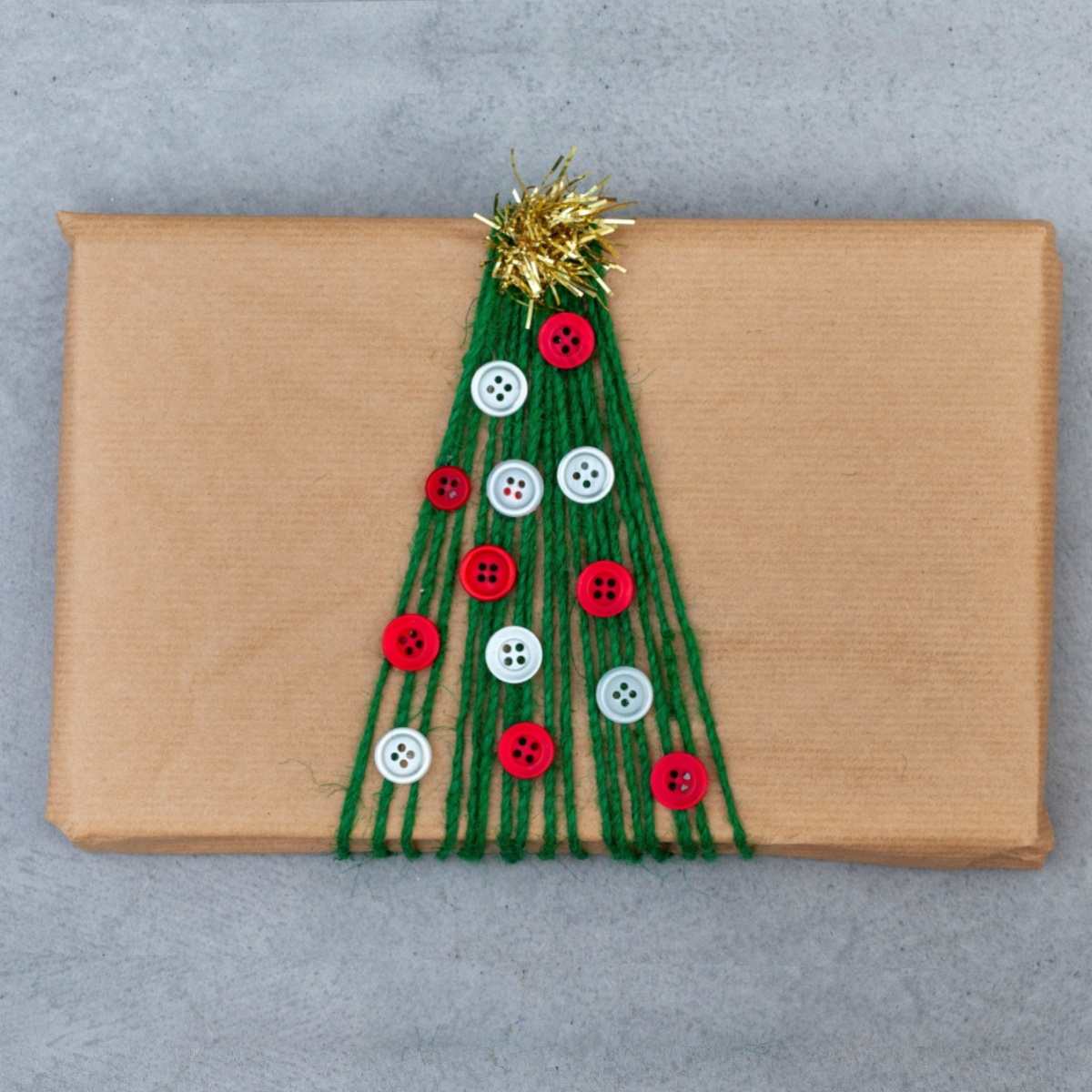 Uma ideia criativa de embalagem de presente de férias mostrando um pacote retangular embrulhado em papel marrom, com fio verde envolvendo-o para se parecer com uma árvore, botões colados para representar enfeites e um pequeno pedaço de guirlanda no topo para se parecer com uma estrela de topo de árvore.