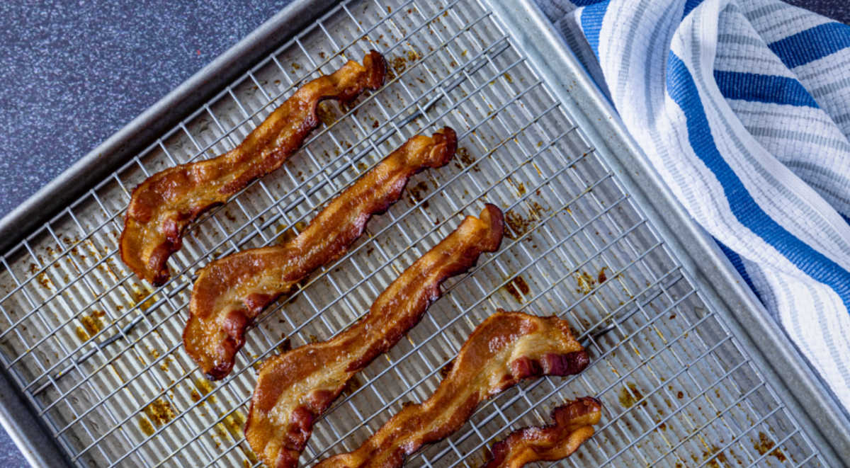 Fatias de bacon crocante cozidas no forno sobre uma grade de arame e assadeira ao lado de uma toalha listrada azul.