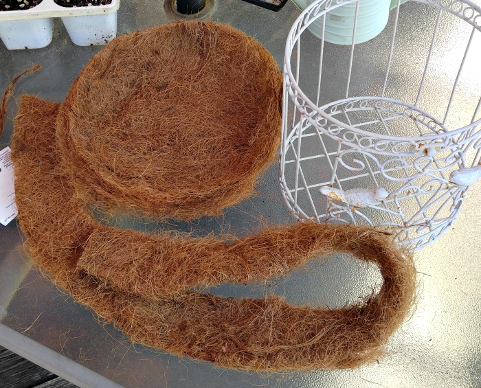 Um forro de fibra de coco cortado sobre uma mesa com algumas pequenas plantas e uma gaiola branca aberta.