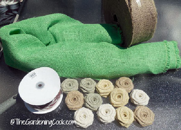 Materiais para fazer uma guirlanda de juta DIY - juta verde, fita com borda de arame e fileiras de flores de rosa de juta.