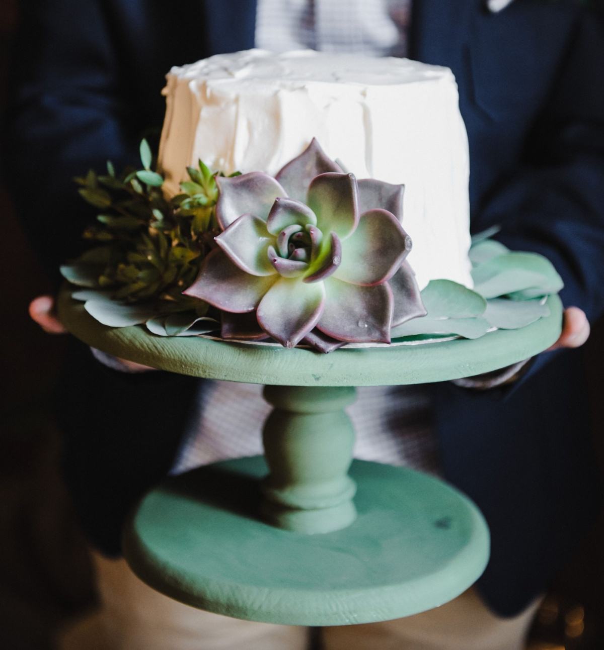 Um bolo com suculentas com cobertura de buttercream branca em um suporte de bolo de madeira verde, com suculentas de pasta de goma ao redor do bolo.