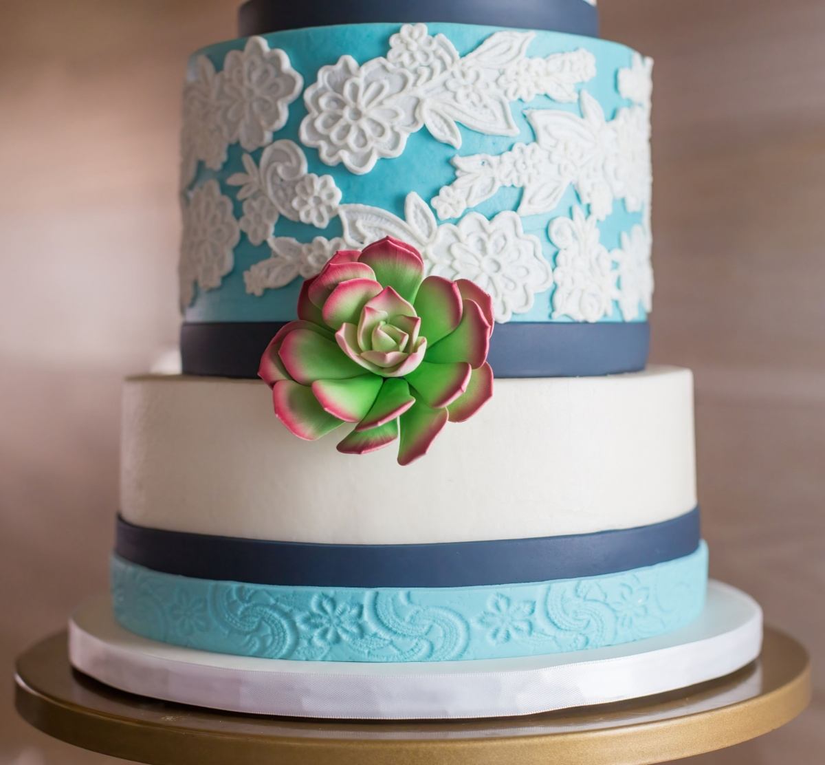 Um bolo de dois andares com suculentas, decorado com fondant branco e azul-tiffany, fita azul-escura e um topo de bolo de suculenta de fondant e pasta de goma no centro.